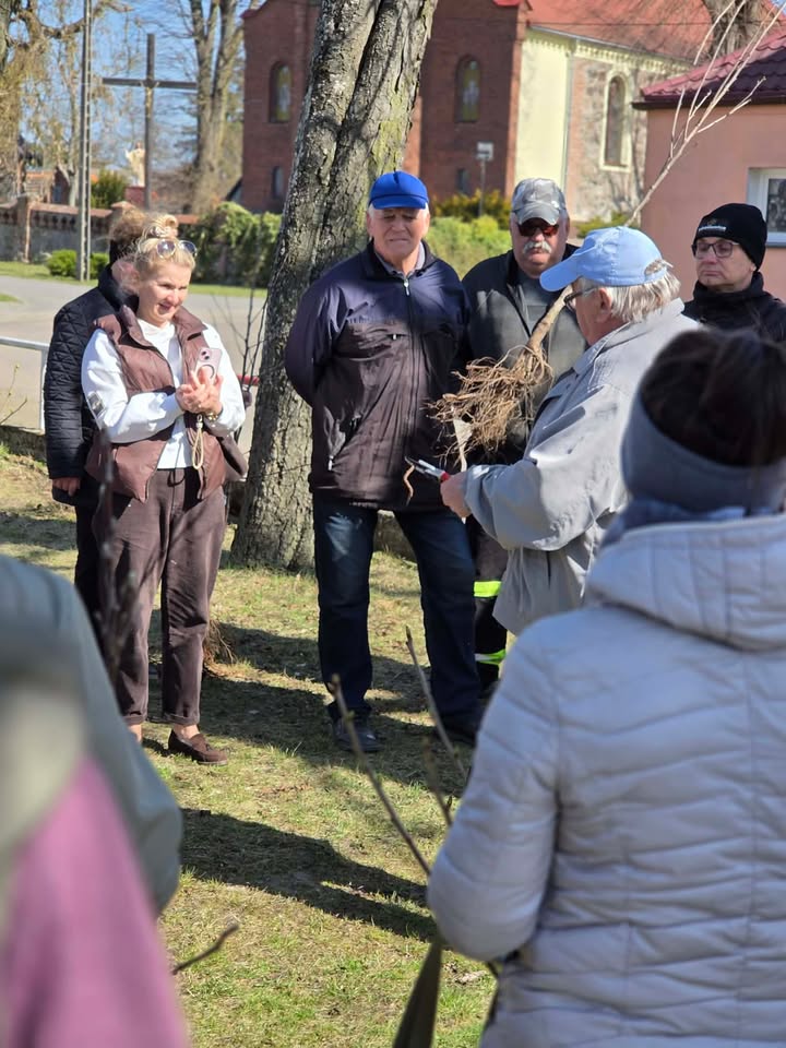 Grupa ludzi stoi na dworze, przed nimi stoi pan trzymjący sadzonkę 