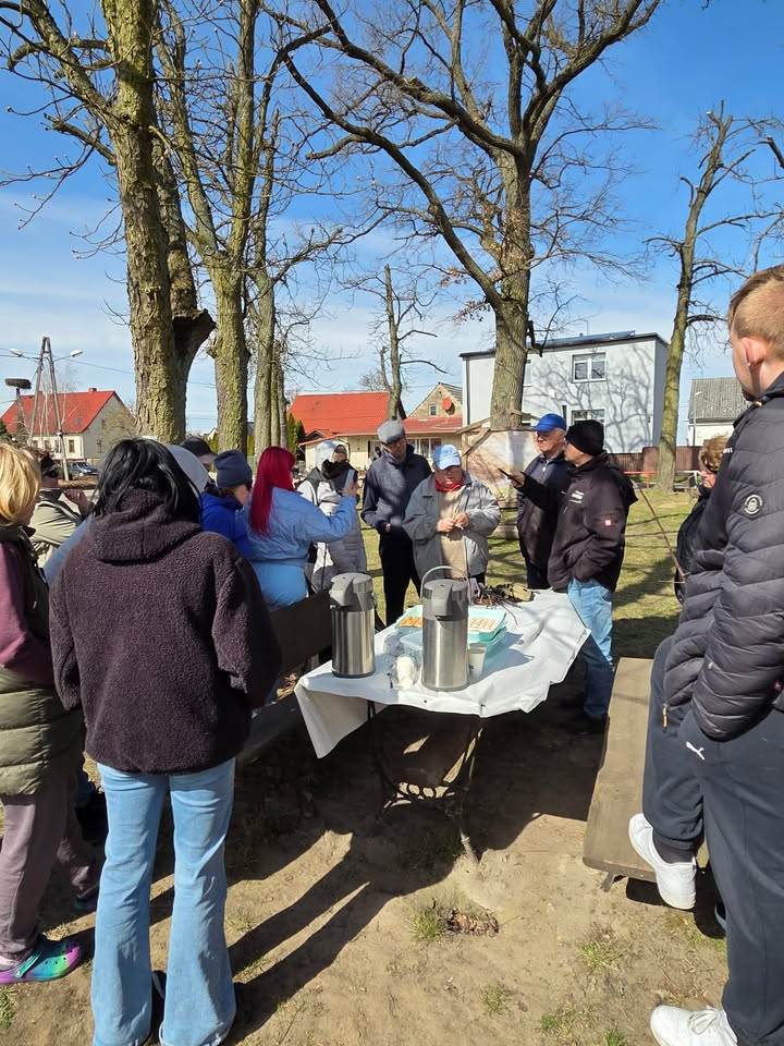 Grupa ludzi stoi na zewnątrz na placu, obok stoi st&oacute;ł, z termosami 