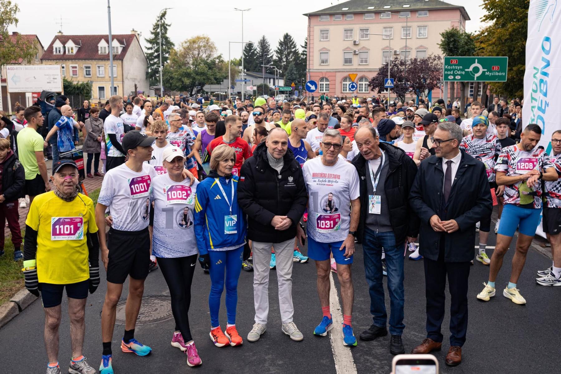 Oficjele stoja na linii startu. Z tyłu grupa biegaczy, budynki