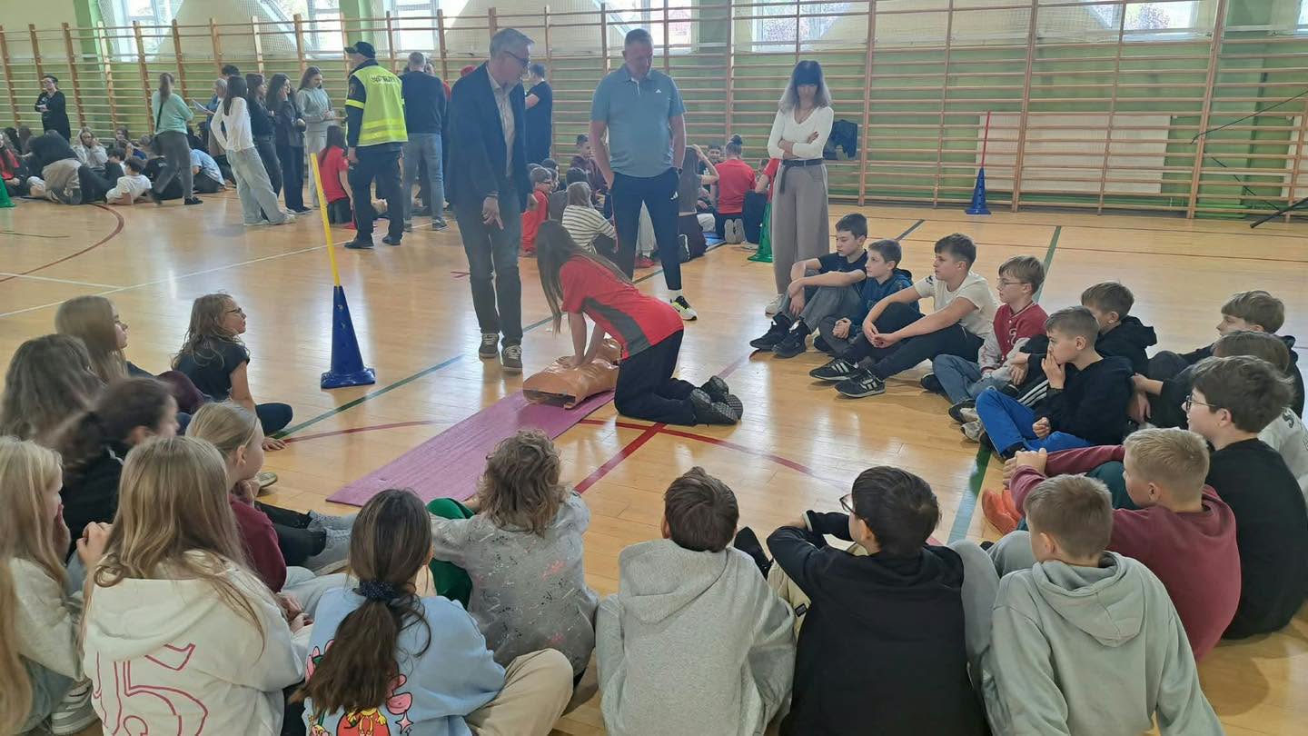 ucnziwoie siedza na podłodze w hali, na środku dziewczyna w czerwonej koszulace na manekinie robi prezentacje, nad nią stoi burmistrz, inny mężczyzna i kobieta