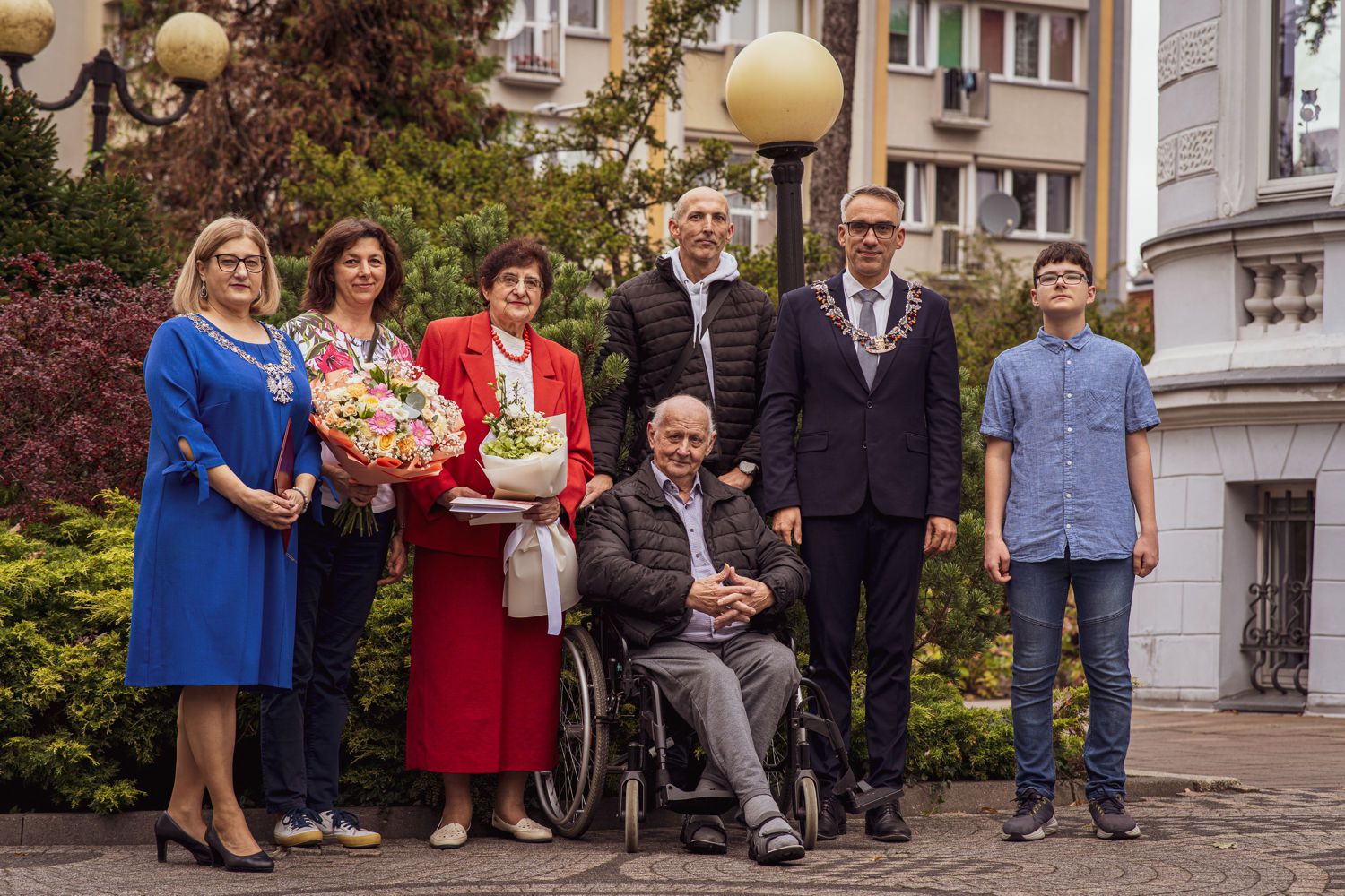 Pamiątkowe zdjęcie plenerowe z okazji jubileuszu. Po lewej stronie stoi Kierownik Urzędu Stanu Cywilnego Maria Zając w niebieskiej sukni z łańcuchem urzędowym, obok kobieta z bukietem kwiat&oacute;w oraz jubilatka w czerwonym żakiecie. W środku siedzi na w&oacute;zku inwalidzkim jubilat, za nim stoi mężczyzna w czarnej kurtce. Po prawej stronie stoi Burmistrz Dębna Wojciech Czepułkowski w garniturze z łańcuchem urzędowym oraz młody chłopiec w niebieskiej koszuli.