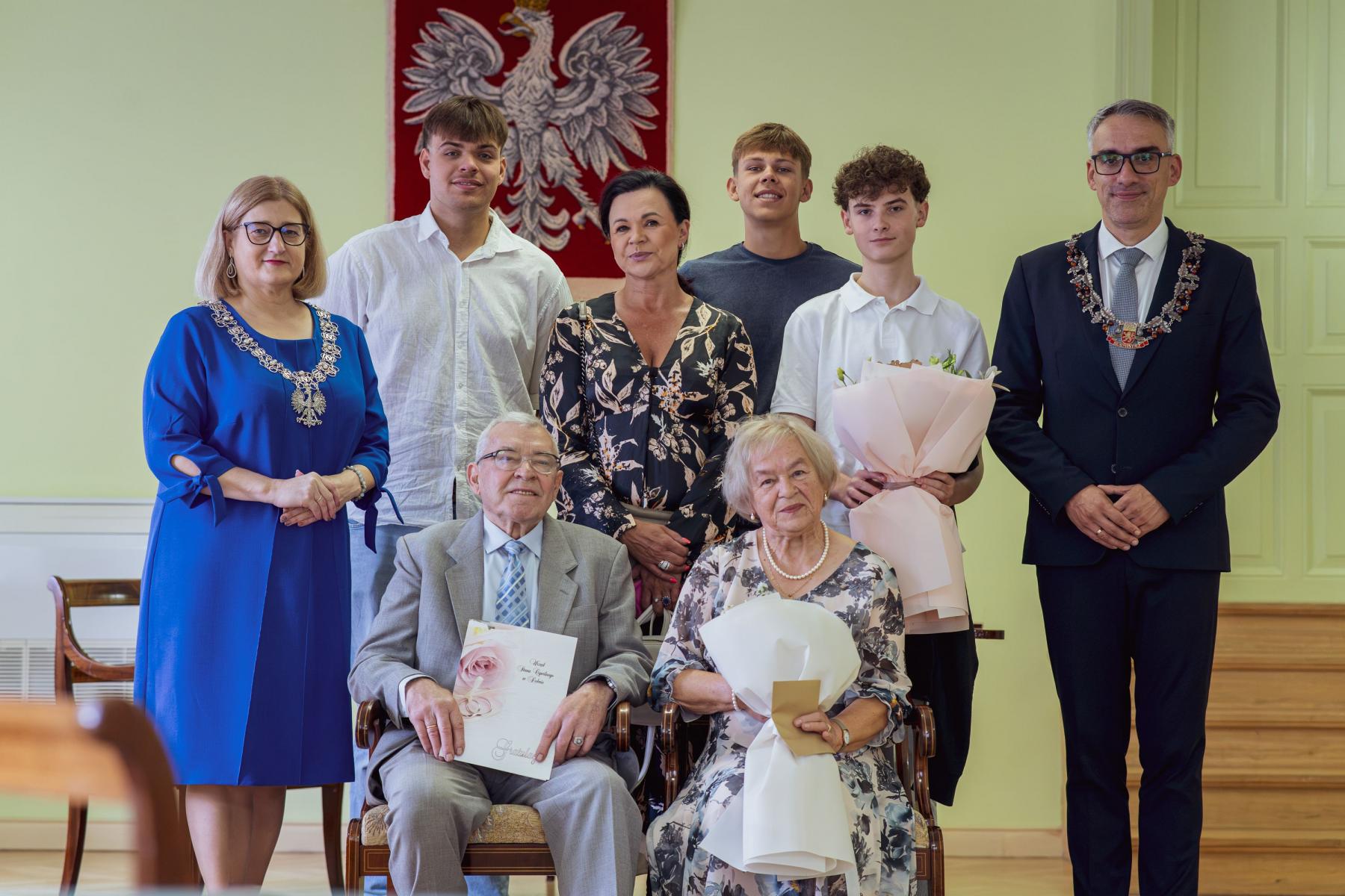 Pozowane zdjęcie grupowe z okazji jubileuszu małżeńskiego we wnętrzu urzędu. Na pierwszym planie siedzą jubilaci: mężczyzna w szarym garniturze trzymający pamiątkowy grawerton oraz kobieta w jasnej wzorzystej sukience z bukietem kwiat&oacute;w. Za nimi stoi rodzina oraz przedstawiciele urzędu. Po lewej stronie stoi Kierownik USC Maria Zając w chabrowej sukni i z łańcuchem urzędowym, a po prawej Burmistrz Dębna Wojciech Czepułkowski w ciemnym garniturze i łańcuchu urzędowym. W tle wisi godło Polski.