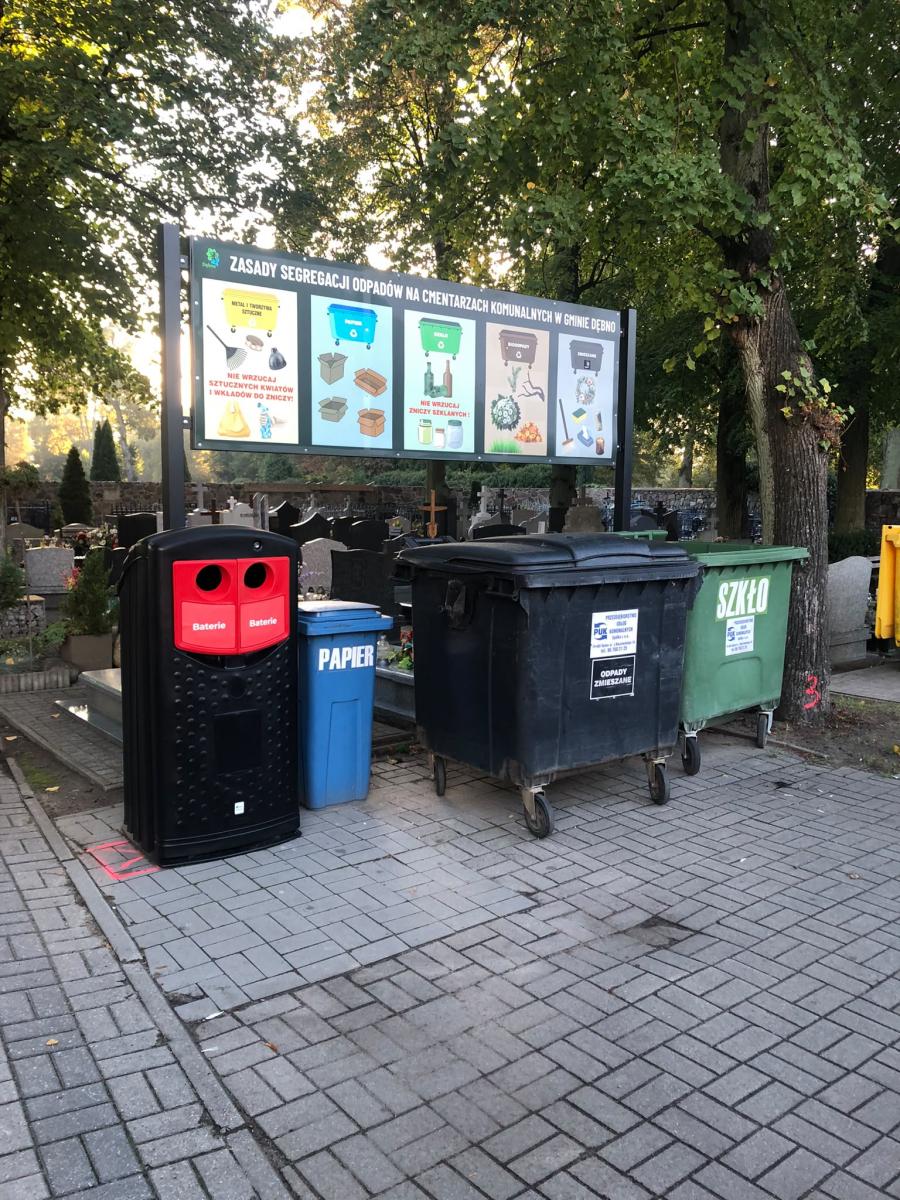 Pojemniki na odpady ustawione w rzędzie. Na baterie pierwszy czarny z czerwonymi wsadami, w tle tablica z zasadami segregacji 