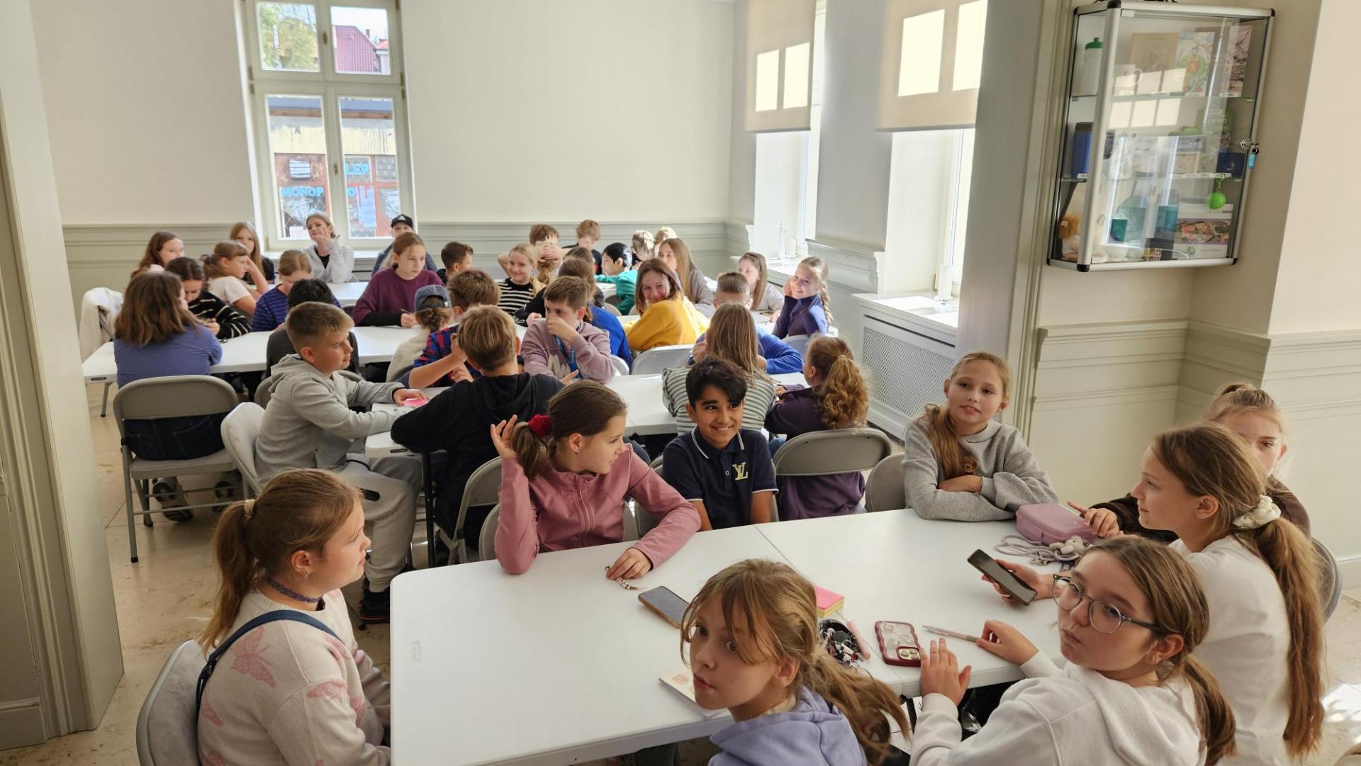 Uczniowie siedzą przy stołach w sali  