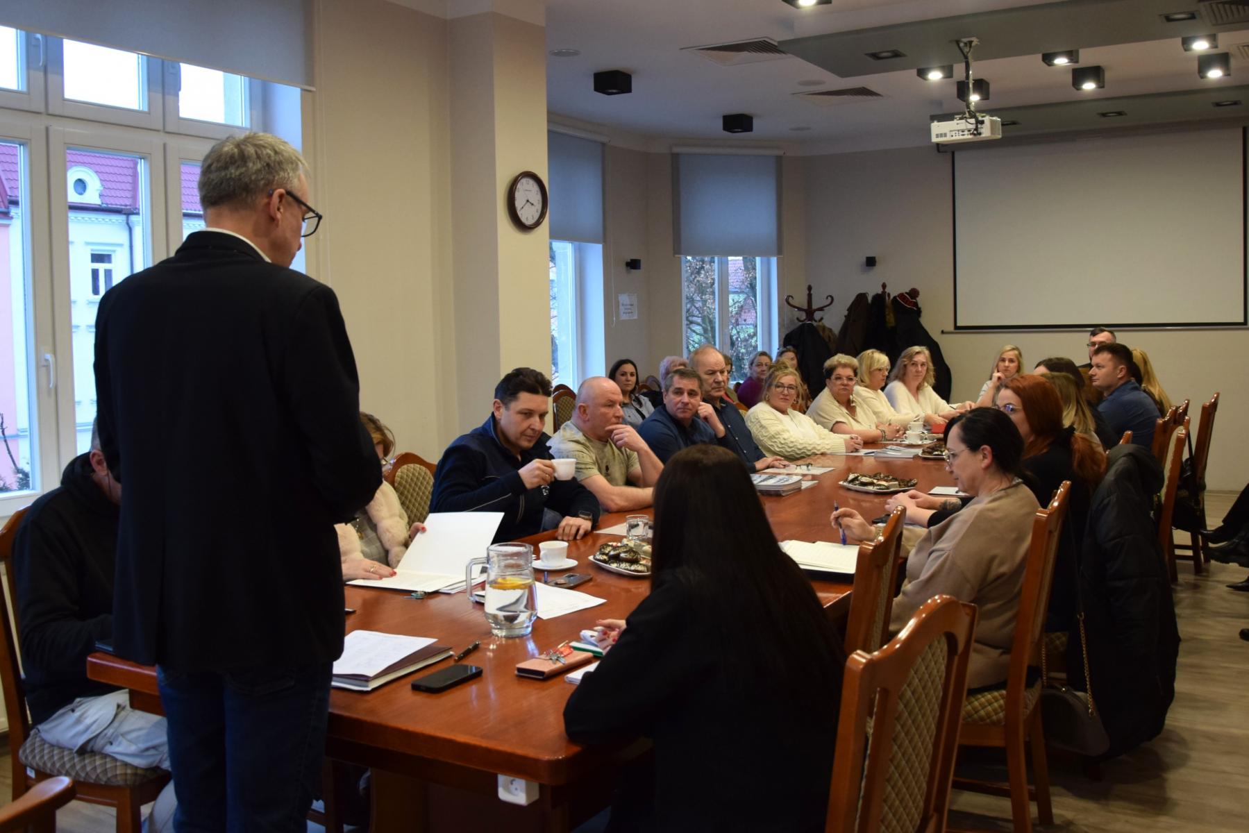 Ludzie siedzą w sali przy stole. Na początku stoi mężczyzna 