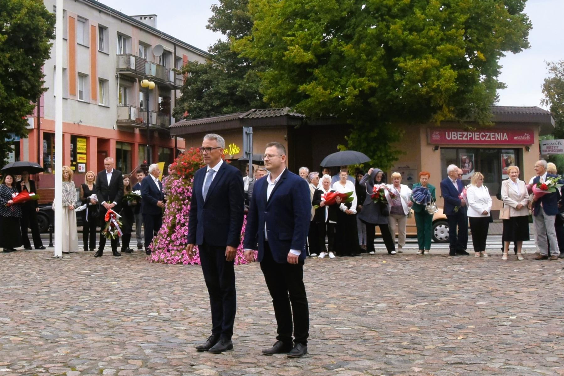 Burmistrz Dębna i radny elegancko ubrani stoją na placu, w tle zgromadzeni uczestnicy 