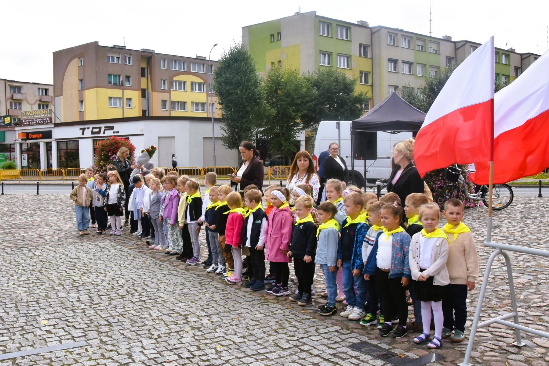 Przedszkolaki stoją w rzędzie na placu, powiewa biało-czerwona flaga 