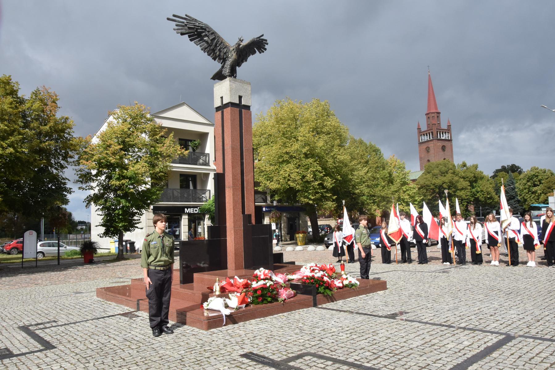 Pomnik Orła Białego, na stopniach leżą biało-czerwone kwiaty, przy pomniku po obu stronach stoją harcerze, w tle drzewa i wieża kościoała 