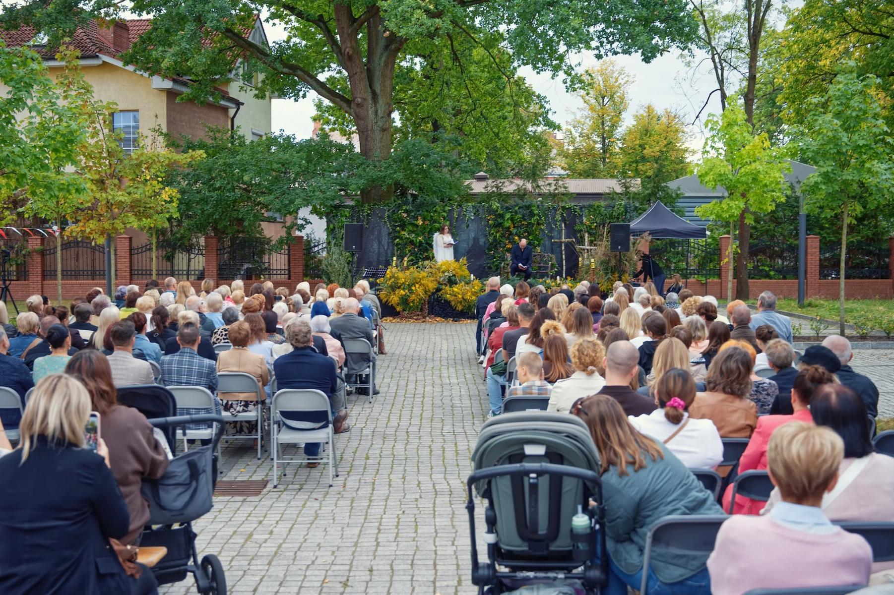 Widok na plac, na kt&oacute;rym na krzesełkach siedzą ludzie, w tle scena w zielonej scenerii, dokoła zielone drzewa, 