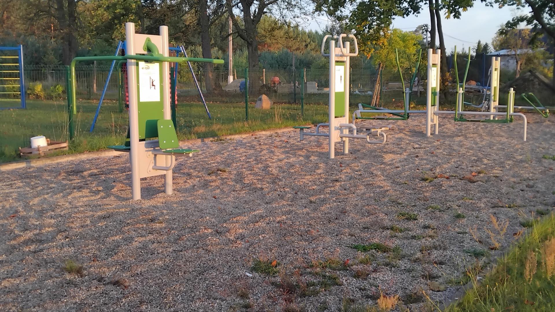 Urządzenia siłowni na nawierzchni żwirowo - piaskowej,  w tle ogrodzenie  i drzewa