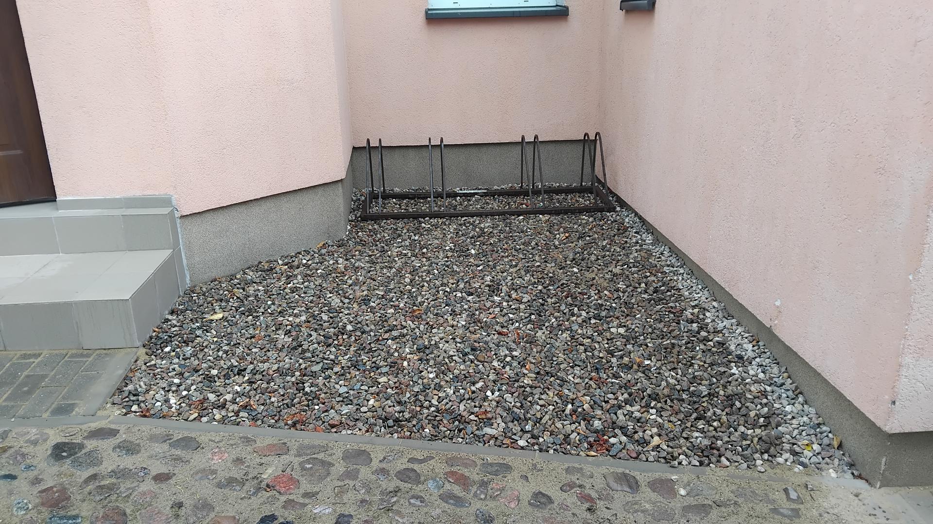 Fragment ściany budynku w Warnicach z nowymi, wyłożonymi szarymi płytkami schodami wejściowymi po lewej stronie. Przy ścianie znajduje się metalowy stojak na rowery, a podłoże wok&oacute;ł niego wyłożono drobnymi kamieniami i żwirem w obramowaniu z kostki.