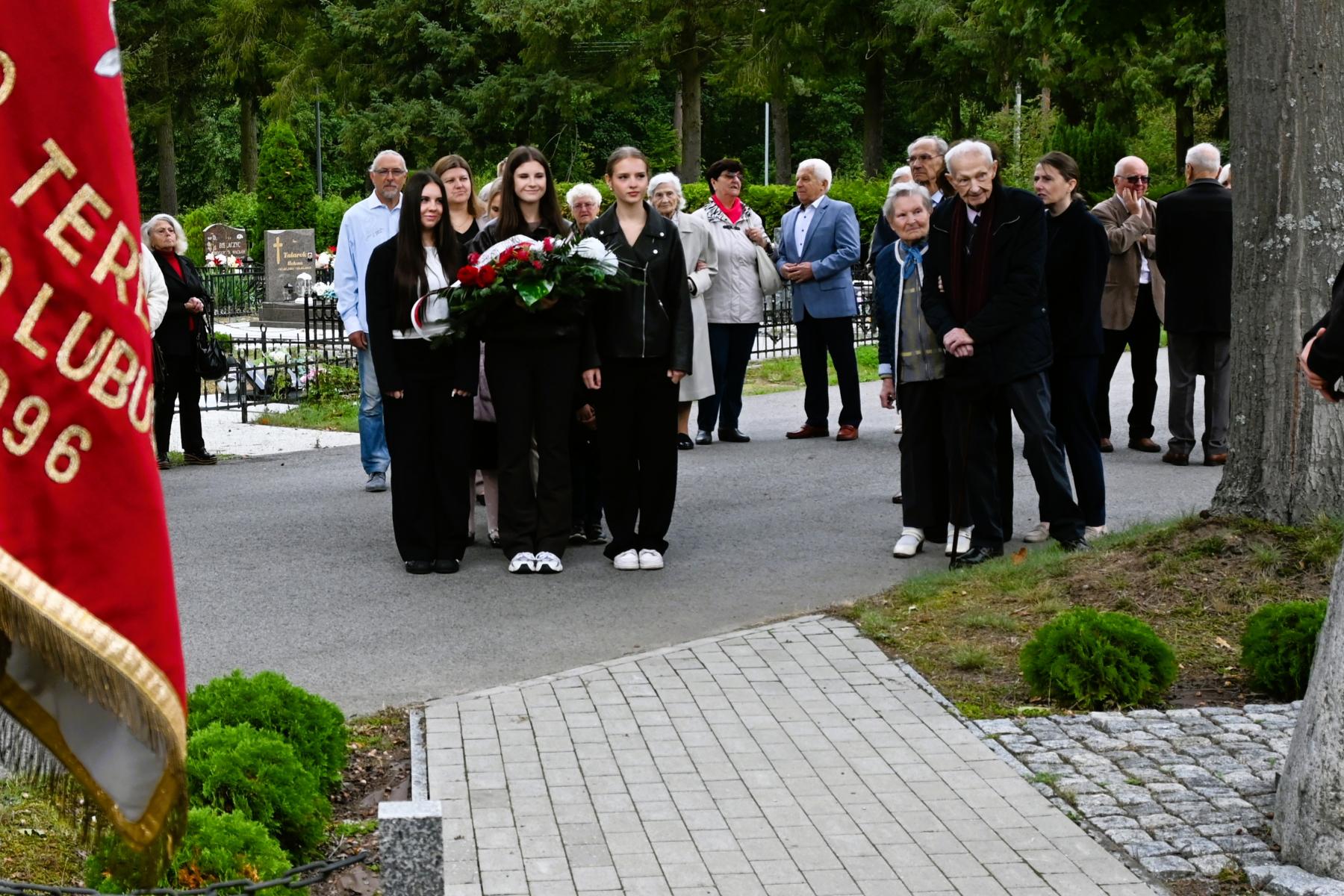 Trzy uczennice liceum (środkowa trzyma wieniec) stoją na alejce wśr&oacute;d innych uczestnik&oacute;w uroczystości 