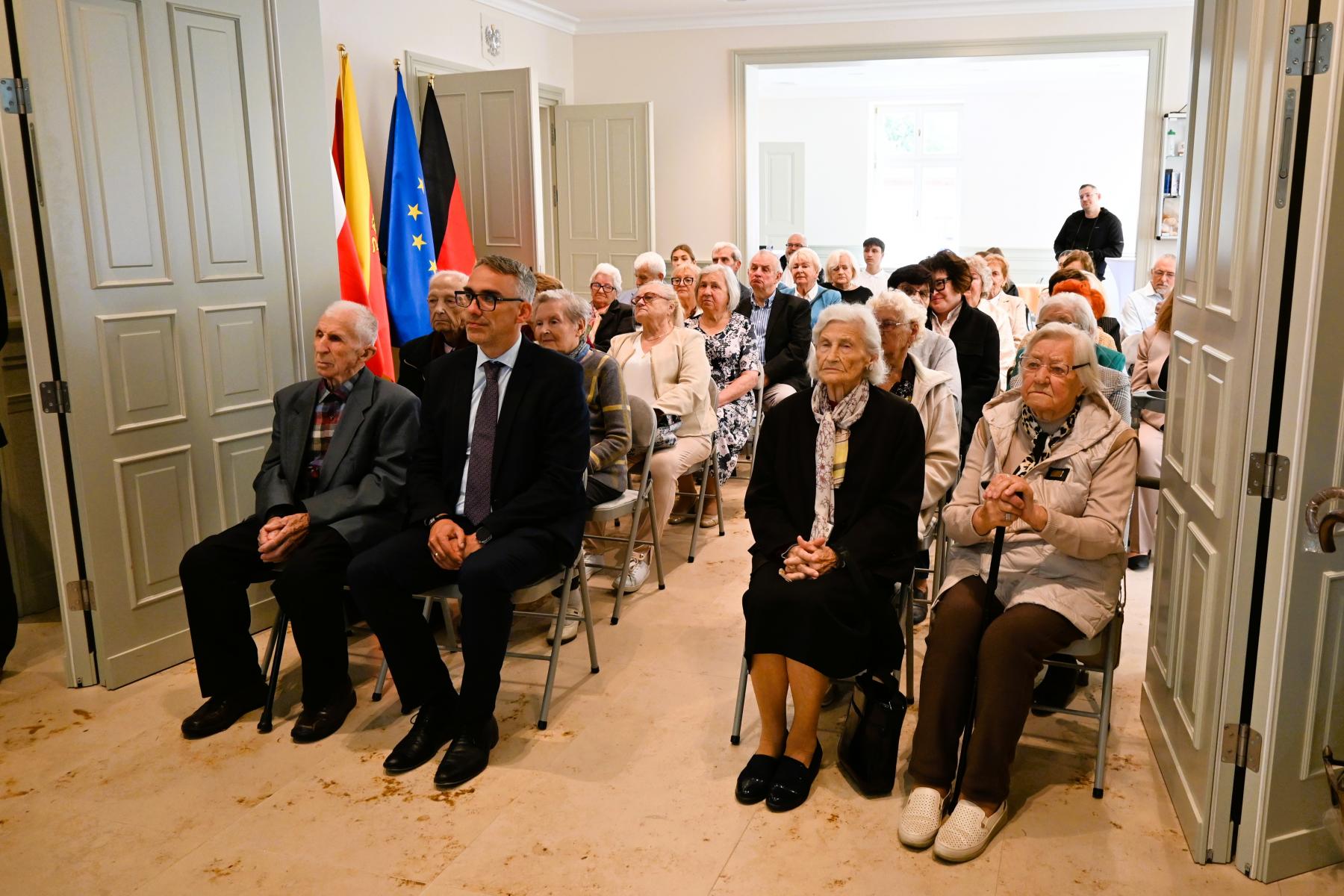 Uczestnicy wydarzenia siedzą w sali w dw&oacute;ch rżedach na krzesełkach. z przodu po lewej siedzi burmistrz Dębna, obok niego sędziwy mężczyzna, po prawej dwie sędziwe panie