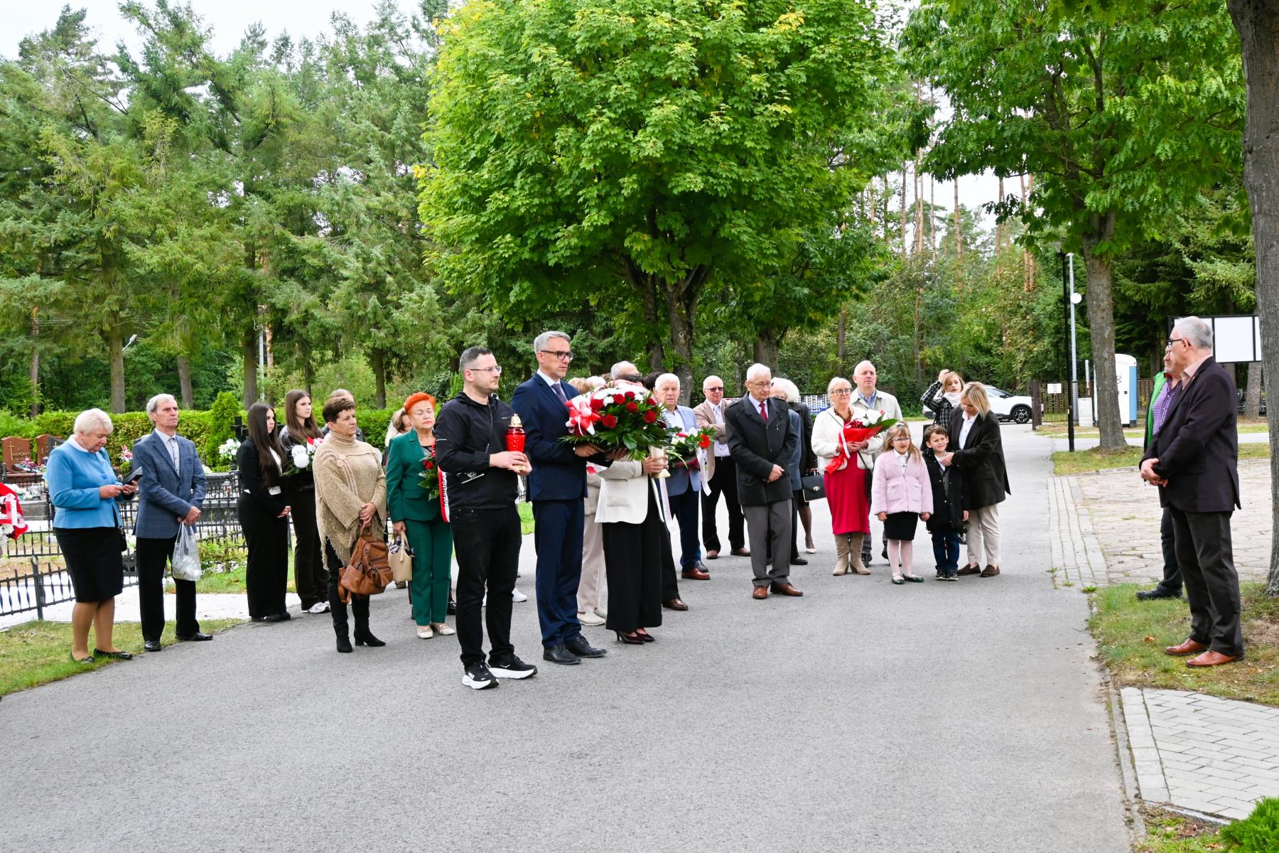 Zgormadzeni uczestnicy stoją na alejce cmentarza, trzymają wieńce 