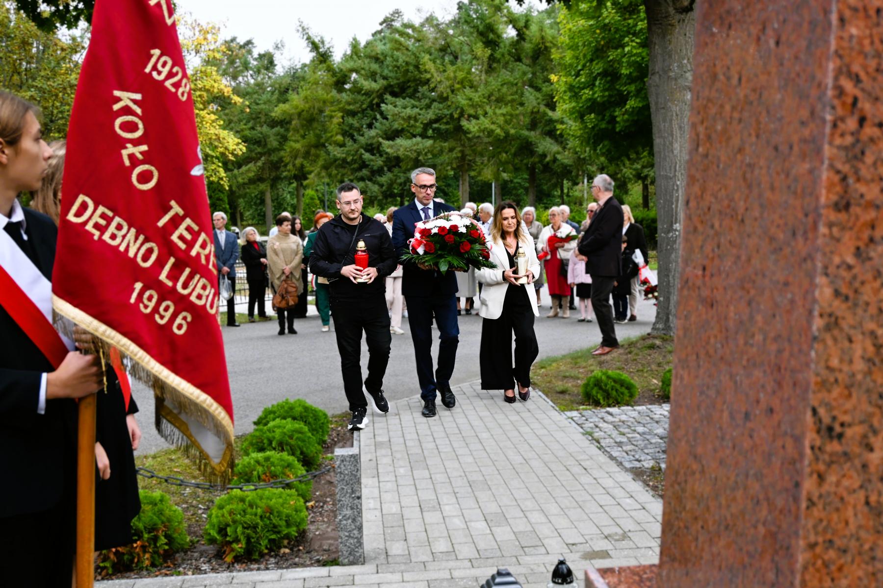 Delegacja trzech os&oacute;b (dw&oacute;ch mężczyzn i kobieta) idą w kierunku pomnika, środkowy niesie biało-czerwony wieniec