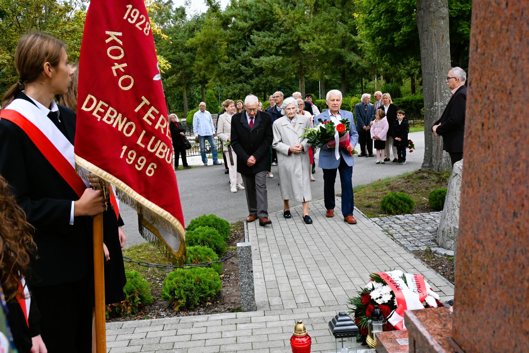Delegacja Sybirak&oacute;w, mężczyzna, kobieta, mężczyna (niesie kwiaty) idą w kierunku pomnika
