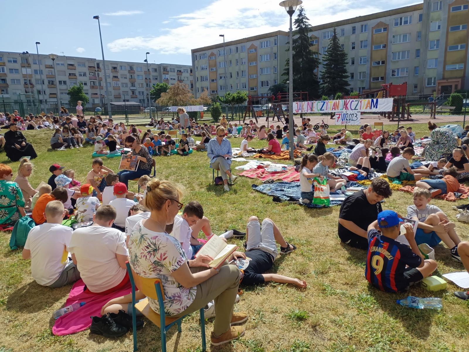 Powiększ zdjęcie. Nauczyciele i uczniwoie siedzą na trawie i czytają książki