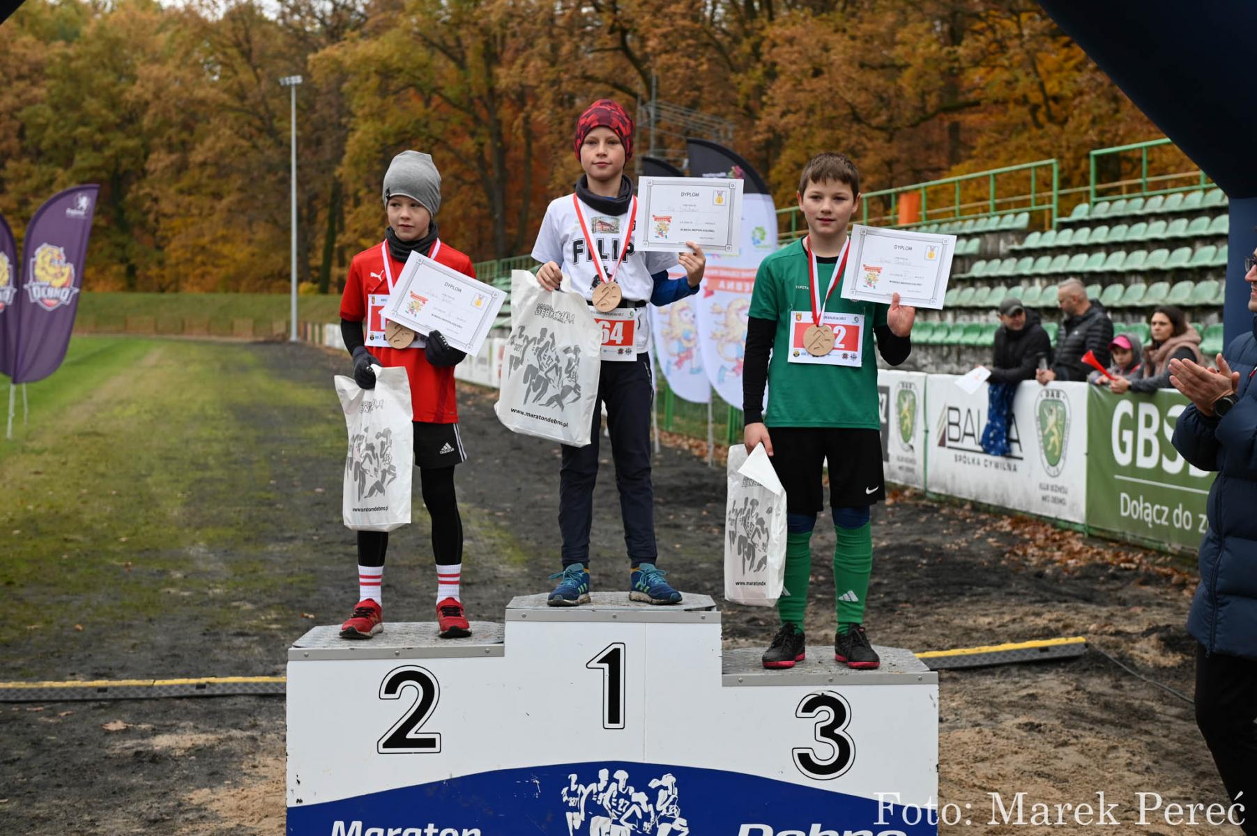 Chłopcy stoją na podium. W rękach trzymają dyplomy. W tle widać siediska stadionowe, drzewa.  