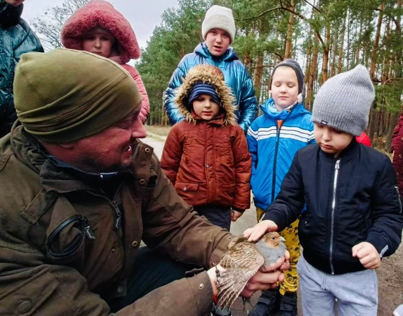 Mężczyzna kuca,  w dłoniach trzyma ptaka, nad nim stoja dzieci i patrzą, jedno dziecko głaszcze
