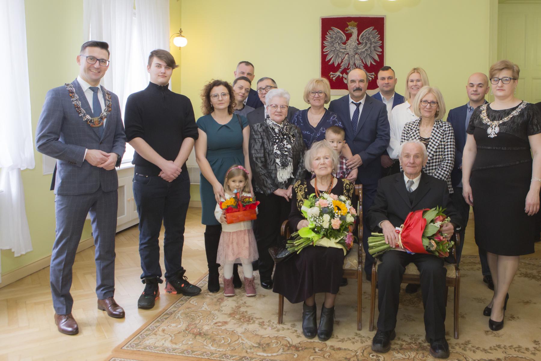 Jubilaci siedzą na krześle, obok stoi rodzina i burmistrz z kierownikiem USC