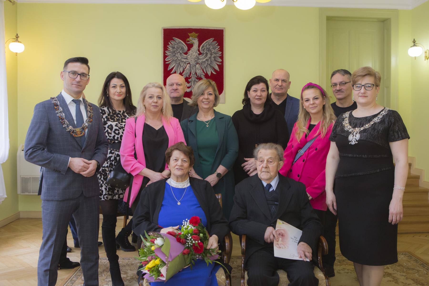 Jubilaci siedzą na krześle, obok stoi rodzina i burmistrz z kierwonikiem USC