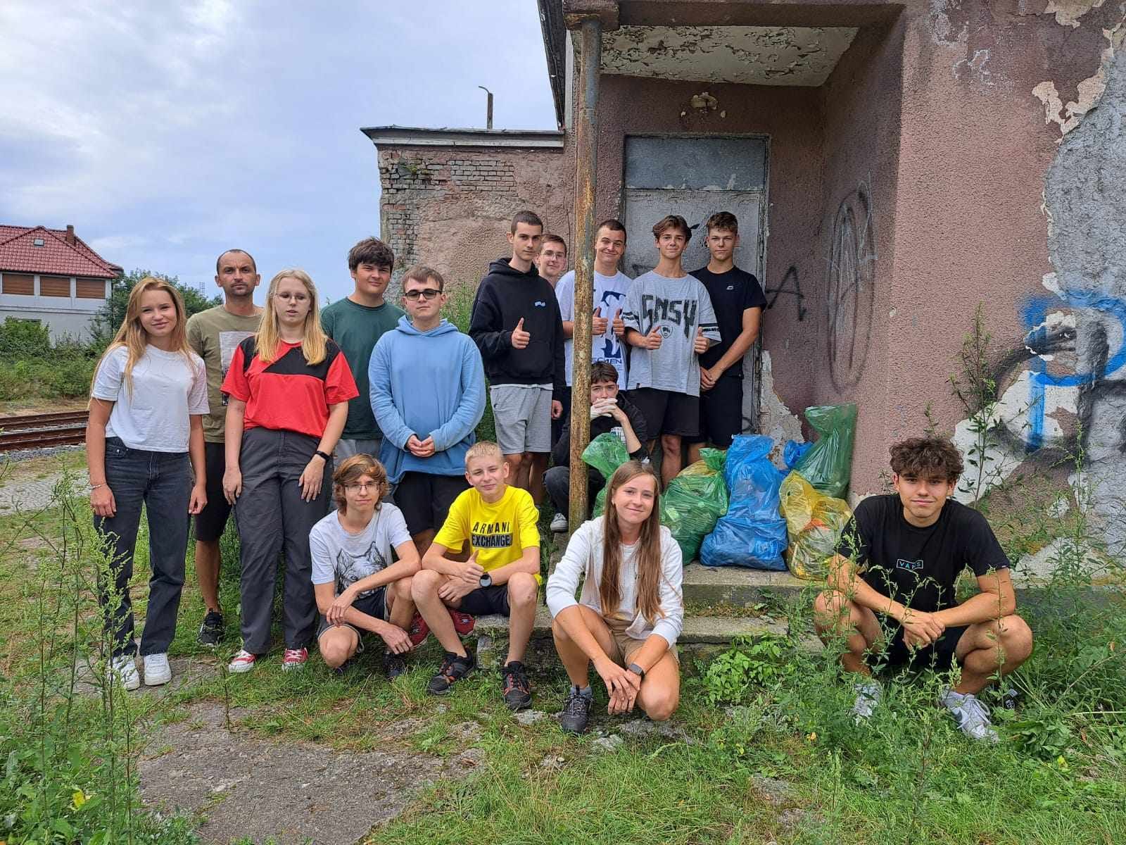 Grupa młodzieży stoi przy starym budynku. Przed nimi pełne worki, obok widać tory