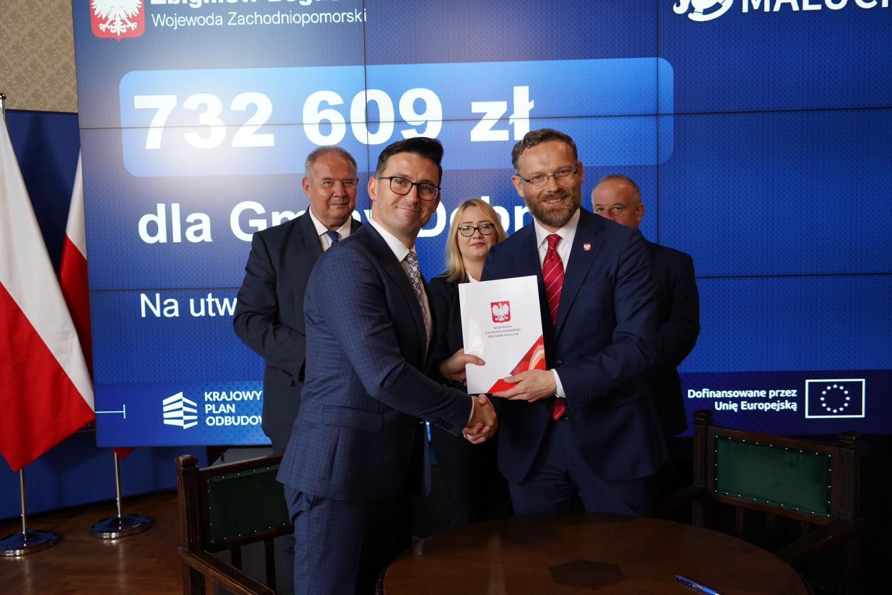 Burmistrz Dębna stoi z wojewodą zachodniopomorskim, w rękach trzymają teczkę z orzełkiem. w tle stoją trzy osoby
