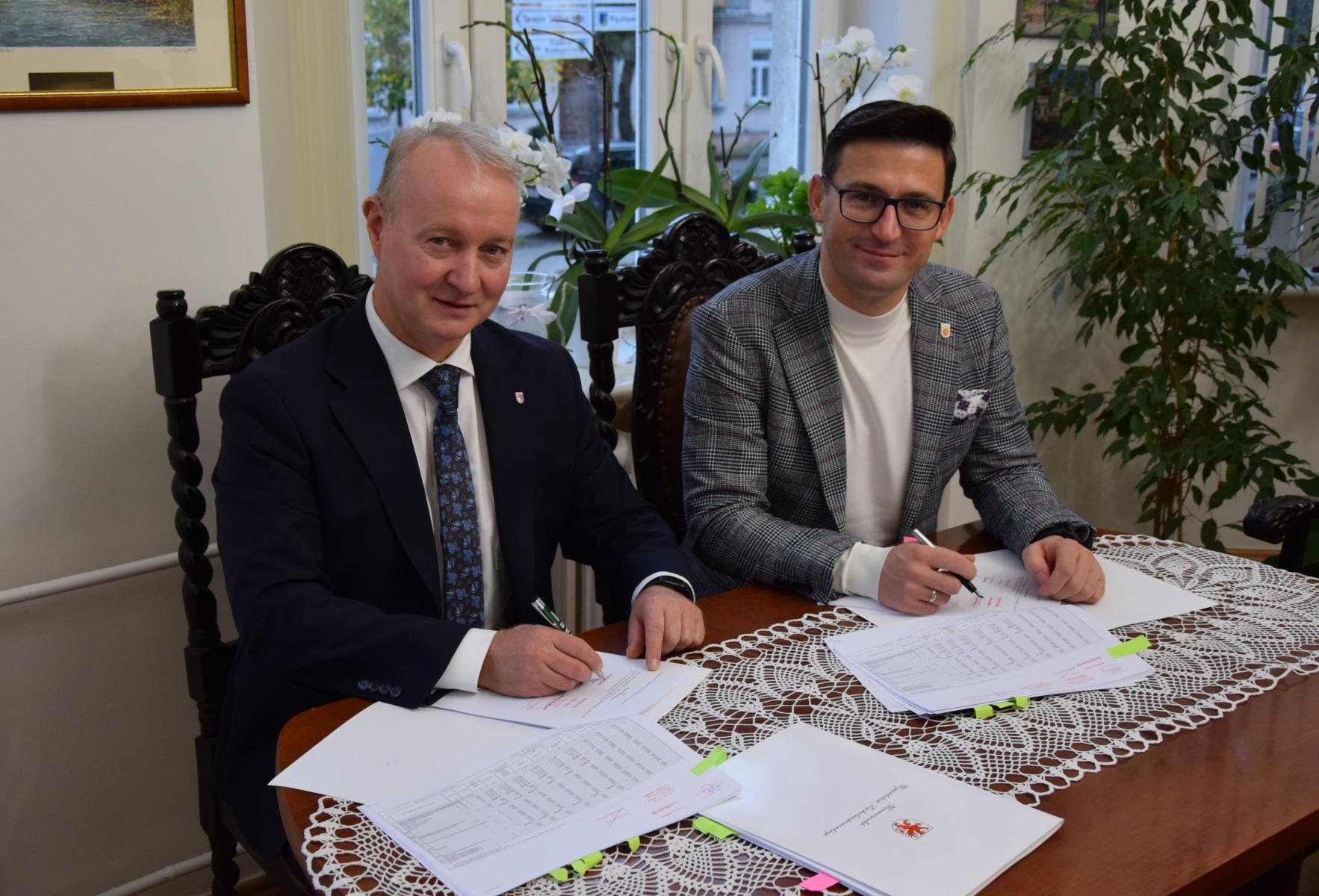 Od lewej: O. Kustosz i G. Kulbicki siedzą przy stoliku, podpisują umowy. W tle na parapecie widac storczyki 