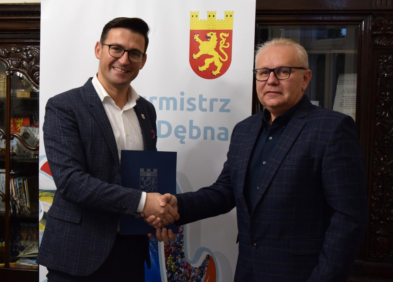 Od lewej stoi burmistrz Dębna, który ściska dłoń prezesowi firmy. Burmistrz uśmiechnięty trzyma teczkę granatową. W tle tło na którym widnieje herb gminy 