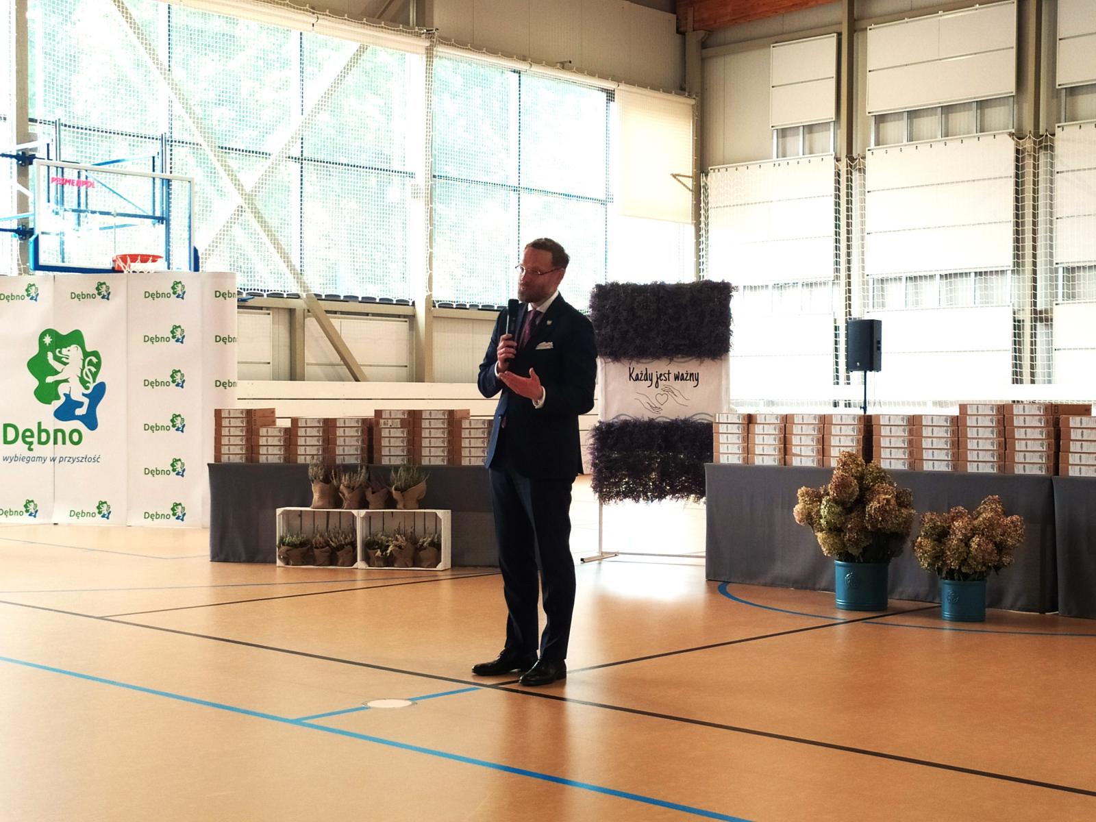Wojewoda stoi na hali sportowej, w ręku trzyma mikrofon, przemawia