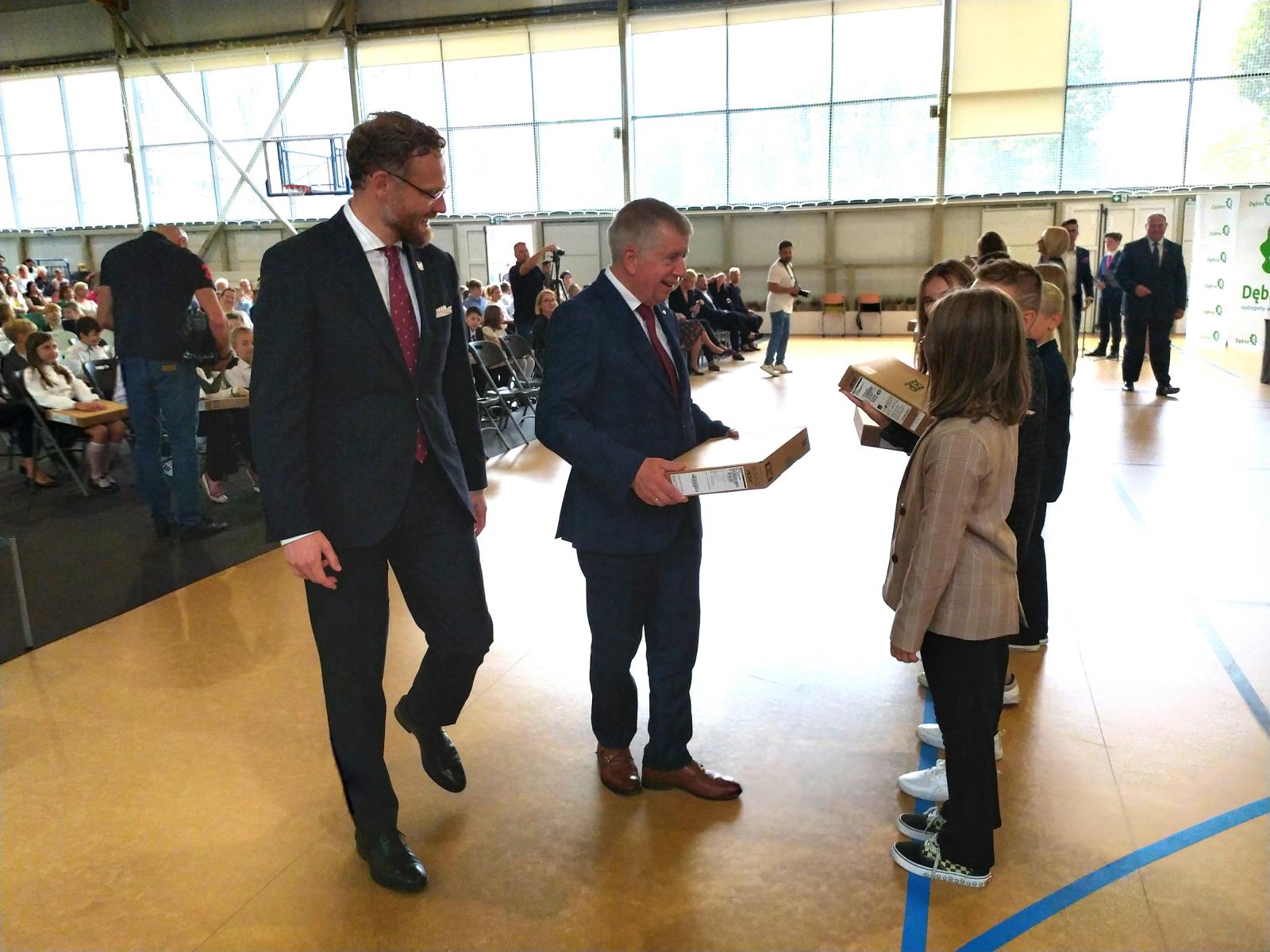 Uczniowie na hali sportowej stoją w rzędzie. W rękach trzymają kartonowe opakowania. 