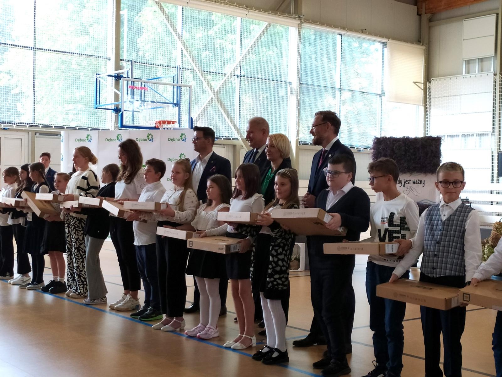 Uczniowie na hali sportowej stoją w rzędzie. W rękach trzymają kartonowe opakowania. 