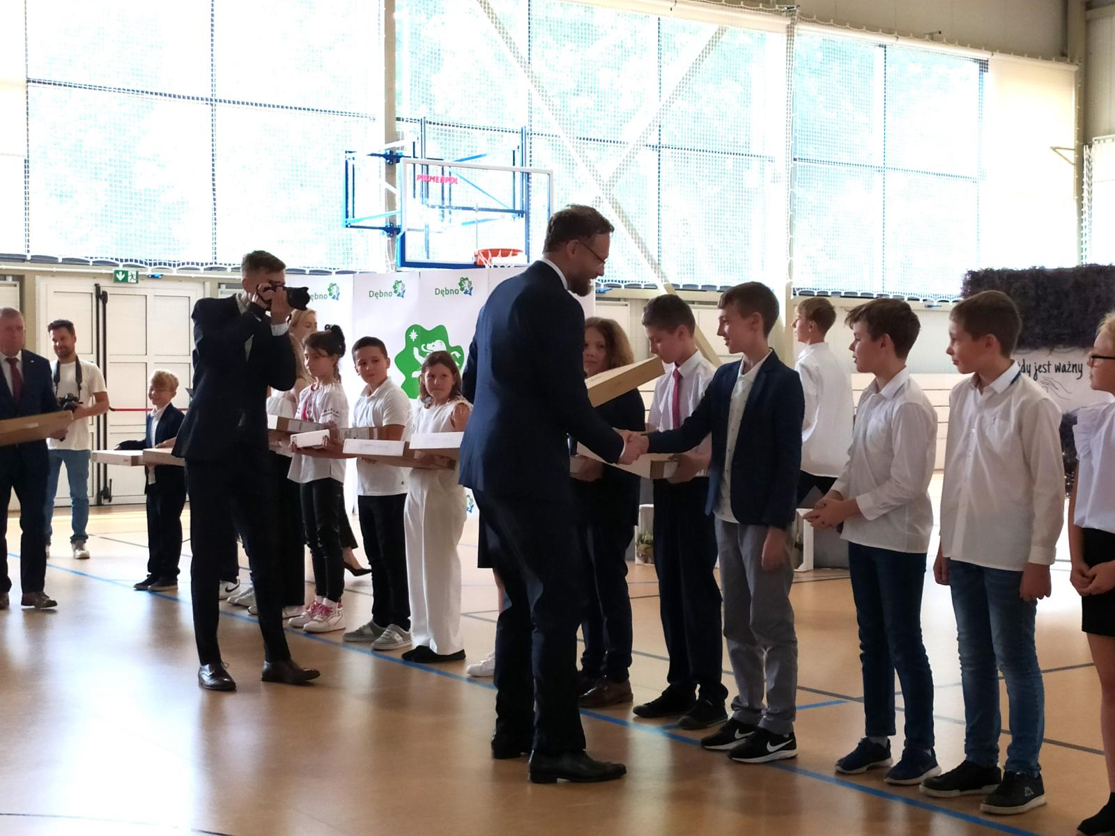 Uczniowie na hali sportowej stoją w rzędzie. W rękach trzymają kartonowe opakowania. 