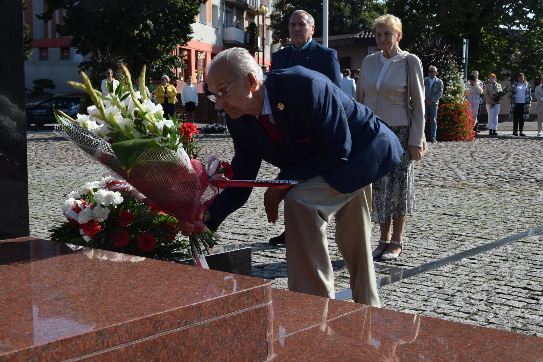 Powiększ zdjęcie. Kwiaty składa przedstawiciel kombatantów, w tle starsza kobieta i mężczyzna 