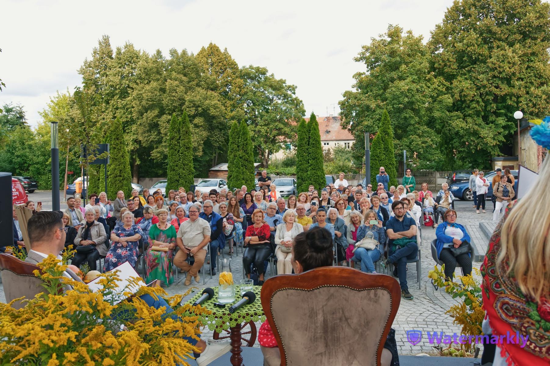 Zgromadzona publiczność siedzi na zewnątrz , w tle krzewy i drzewa. Na pierwszym planie tyłem beżowych fotelach siedzą aktorka i burmistrz