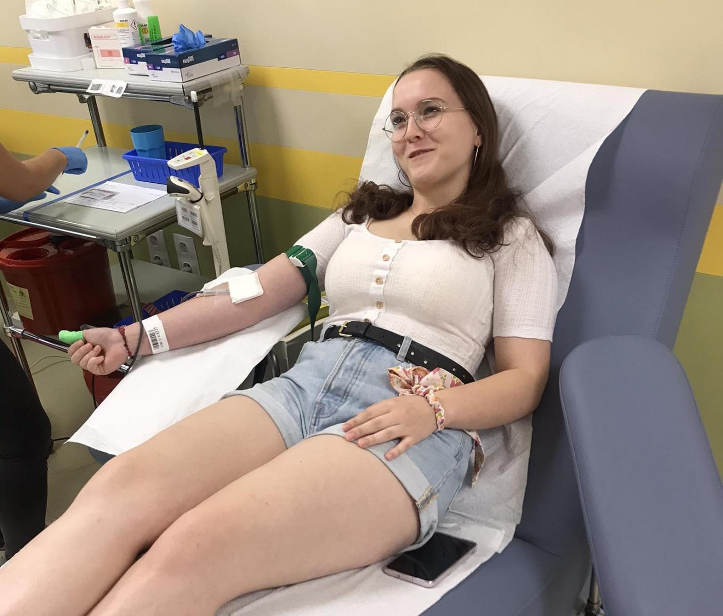 Powiększ zdjęcie. Młoda uśmiechnięta dziewczyna w pozycji półleżącej na fotelu oddaje krew. Z prawej strony stolik z różnymi przyrządami