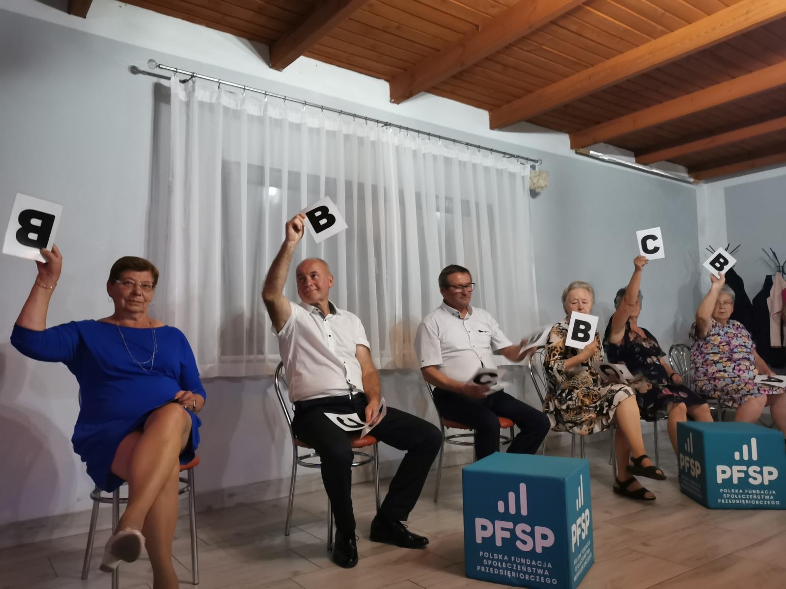 Sześć osób siedzi na krzesłach,. Każdy w dłoni ma podniesioną kartke, 5 z literką B, jedna z C, w tle okno z białą firanką, przed nimi dwie większe kostki z napisem PFSP