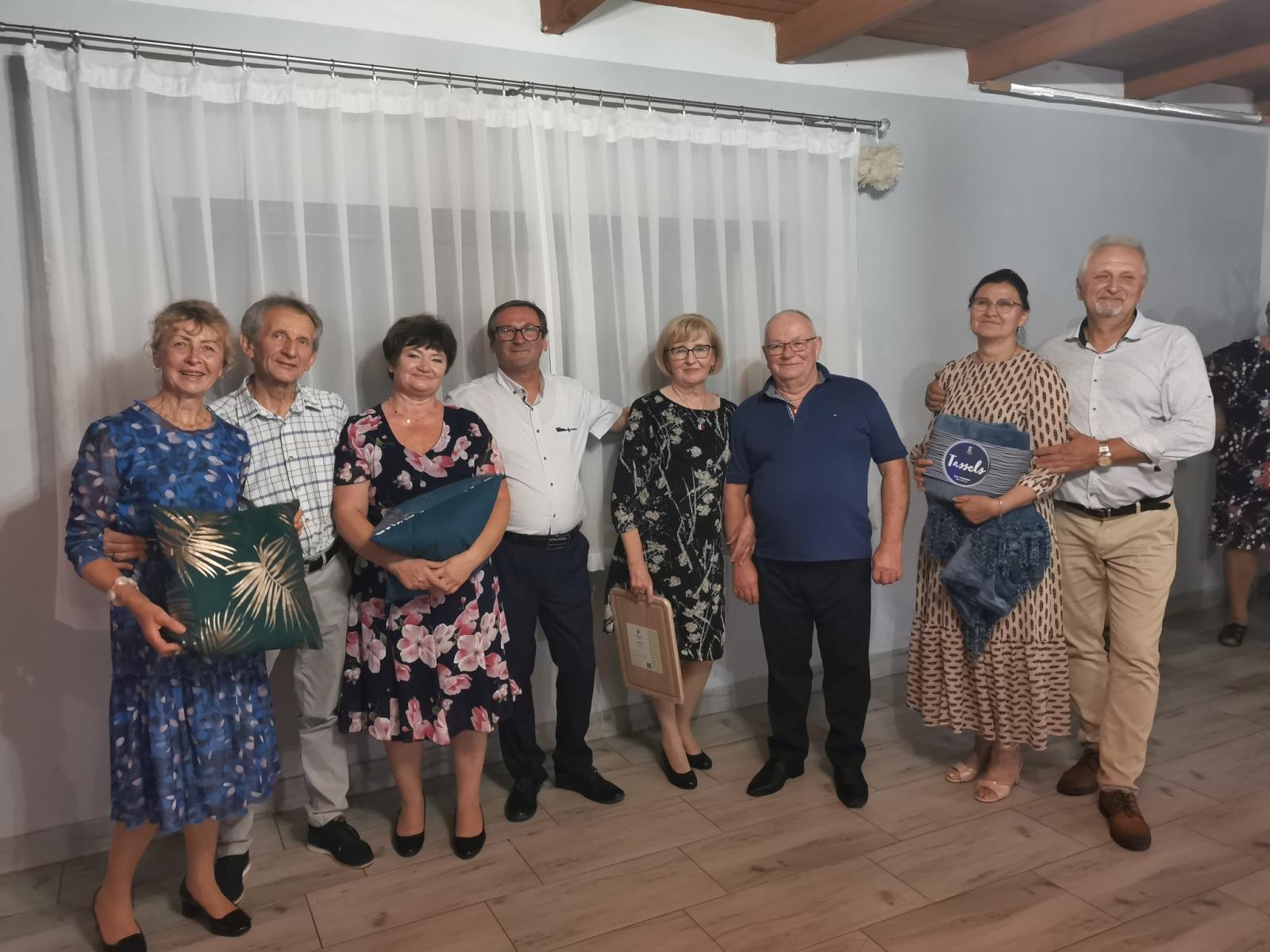 Grupa starszych ludzi stoi w rzędzie. w dłoniach trzymają różne nagrody. w tle okno z białą firaną 