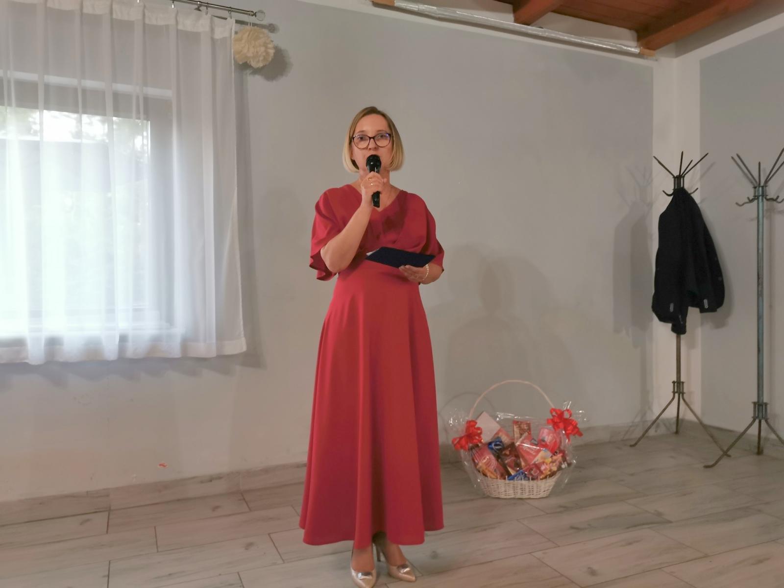 Powiększ zdjęcie. Pani wiceburmistrz stoi, trzyma mikrofon w dłoni przy ustach, ubrana w długa czerwoną suknię, w tle na ziemi stoi kosz z kokardami, za nia okno z białą firanką