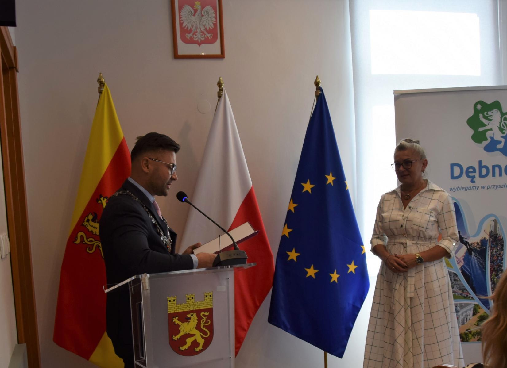 Od lewej przy mównicy stoi przewodniczący Rady, ubrany w garnitur, po prawej kobieta w jasnej sukni Edyta Gładysz. W tle flaga gminy, Polski, UE