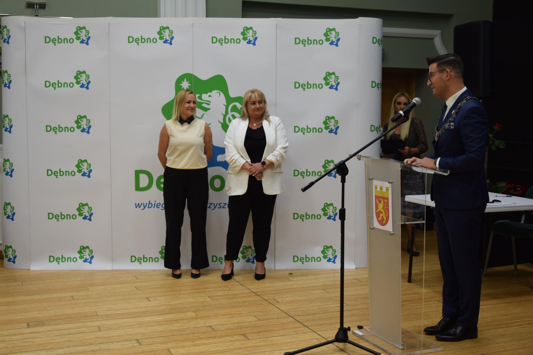 Dwie nauczycielki stoją na tle białej ścianki, na naiej zielono-niebieksie logo i napis Dębno. po prawej stronie przy mównicy przez mikrofon mówi burmistrz Dębna 