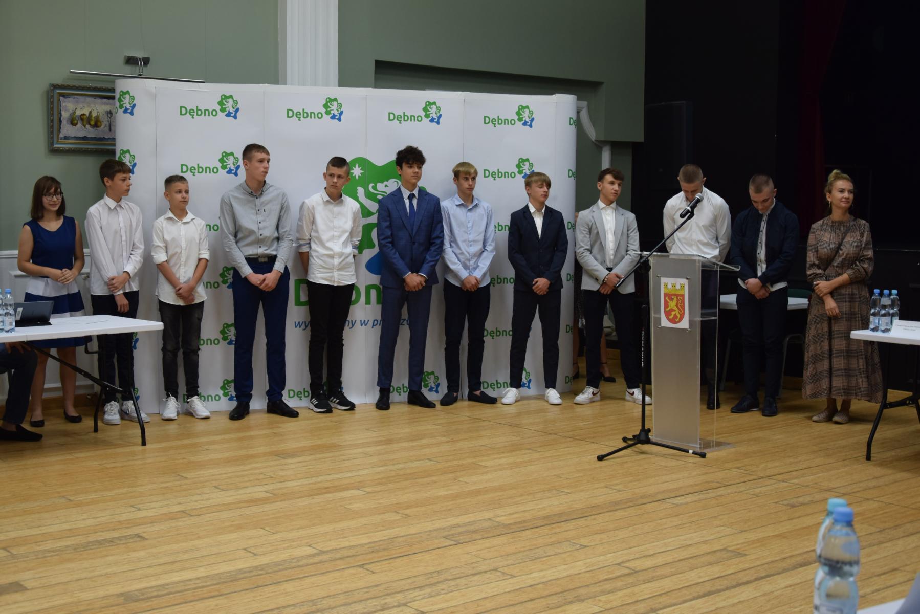 12 uczniów i uczennic stoi w rzędzie, ubrani apelowo, w tle biała ścianka z zielono-niebieskim  logo z napisem Dębno 