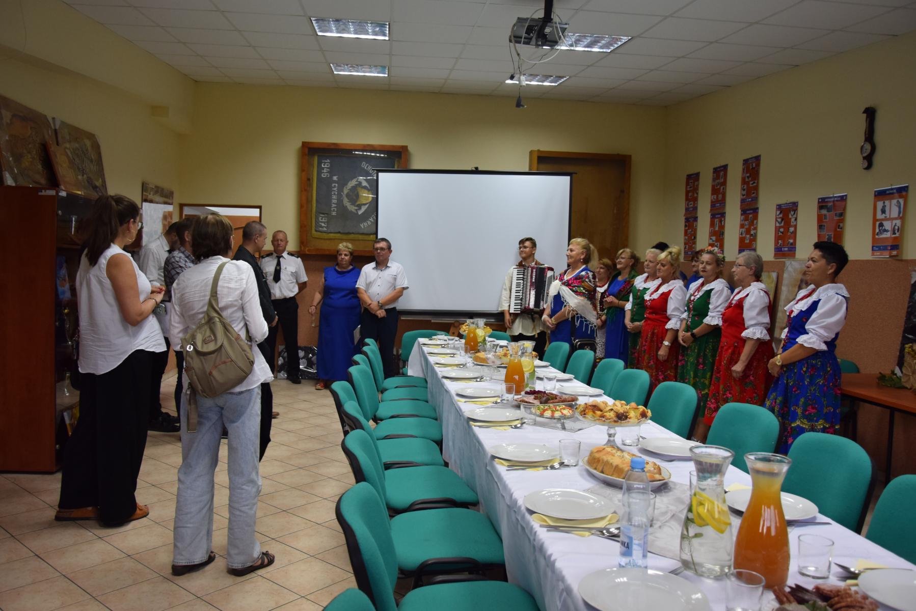 W sali po prawej w rzedzie stoją kobiety w sukniach czerwonych i niebieskich, na końcu młody pan z akordeonem, po srodku biały ekran, po lewej czlonkowie komisji, pani sołtys w nibieskiej długiej sukience, były sołtys