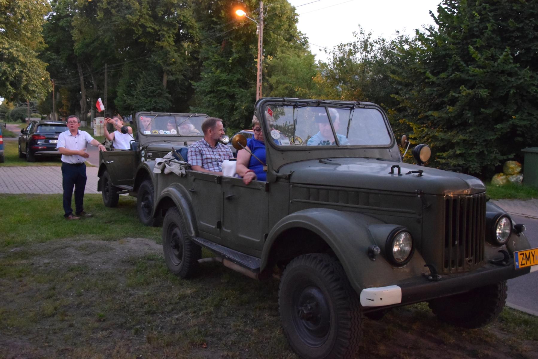dwa samochody wojkowe GAZ, w pierwszym siedzą mężczyźni i kobieta, przy drugim stoi mężczyzna, w środku siedzą osoby.