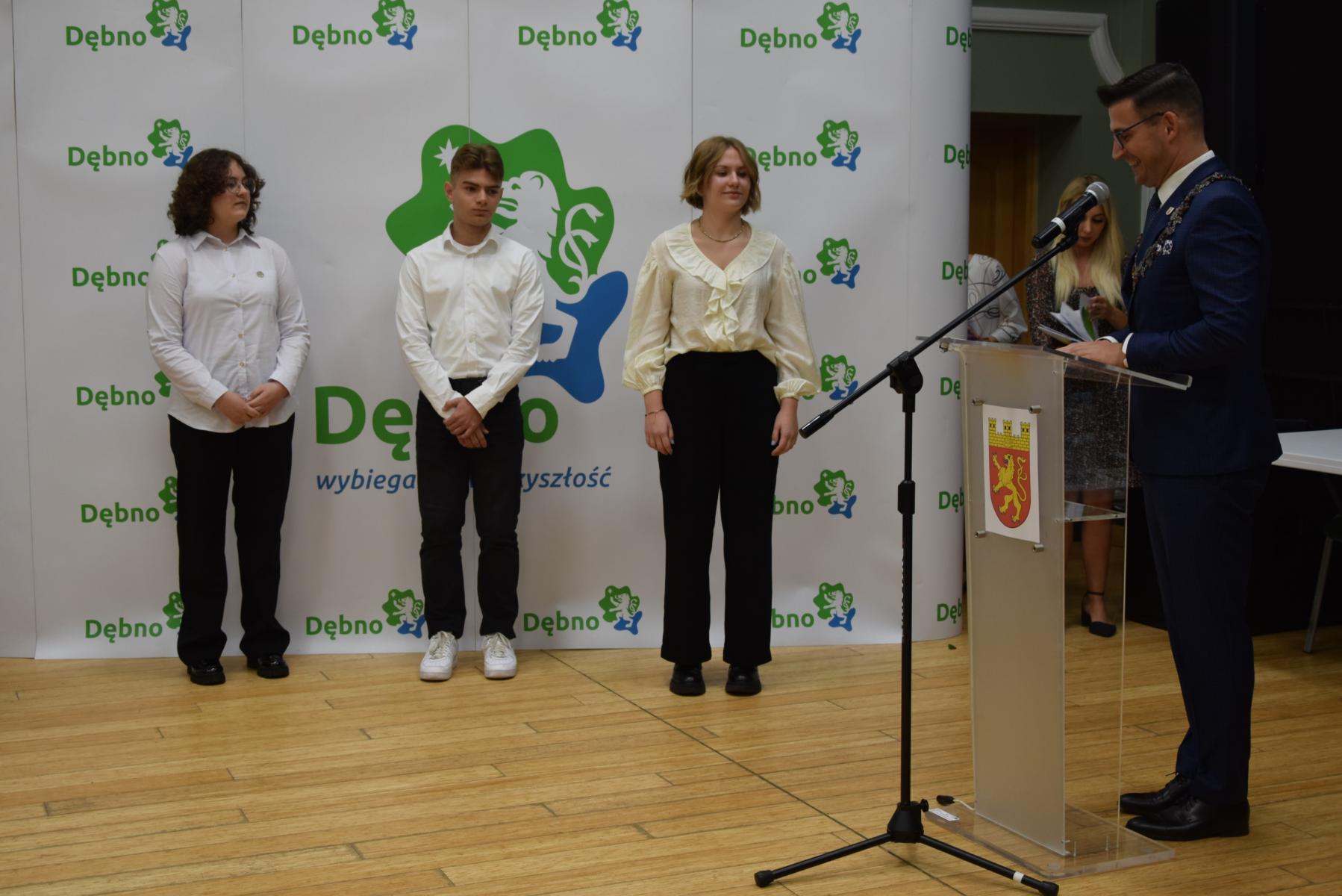 Troje uczniów, ubranych apelowo, stoją w rzędzie na tle białej ścianki. Na niej zielono -niebieskie loga z rysunkowym lwem i napisem Dębno. Z prawej strony stoi burmistrz przy mównicy, mówi przez mikrofon