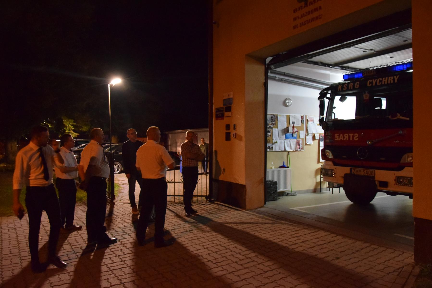 Strażak Ochotnik pokazuje komisji wóz strażacki, który stoi w remizie, w tle pali się lampa, zmierzch