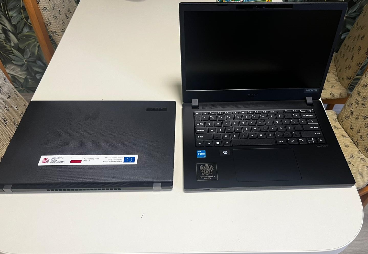 Powiększ zdjęcie. Dwa czarne laptopy leżą onok siebie na białym stole. Jeden zamkniety z prostokątną naklejką z logami, drugi otwarty, na dole w rogu znaczek orła