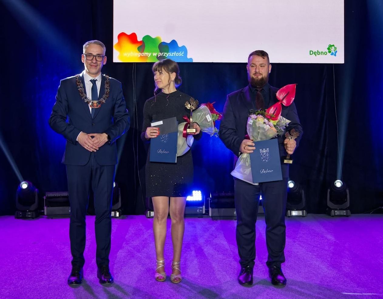 Od lewej elegancko ubrani stoją: burmistrz Dębna, Aneta Dobropolska, Paweł Herbin  (w rękach trzymają statuetkę, kwiaty i teczkę)